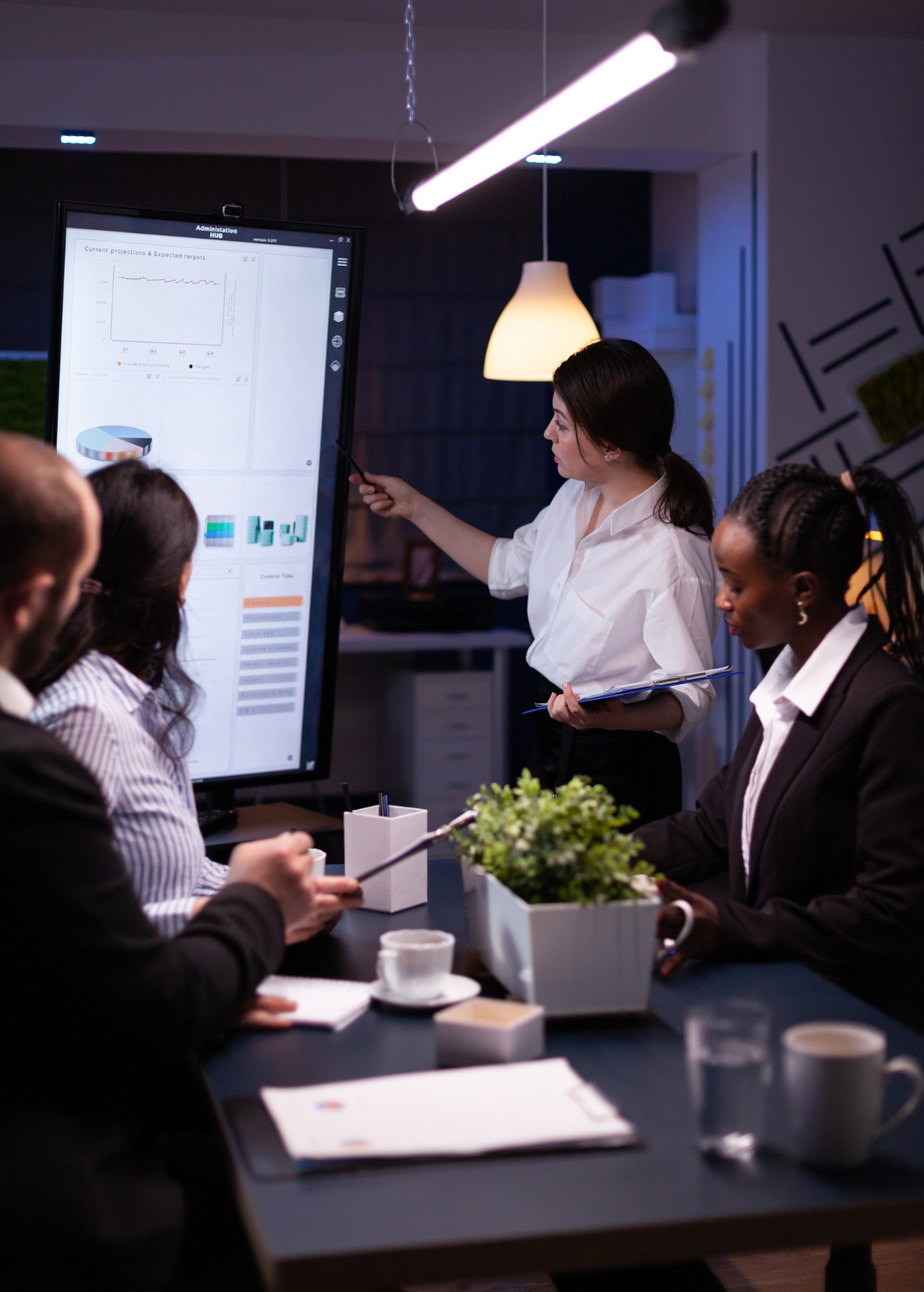 Woman presenting charts on digital screen to other people around table